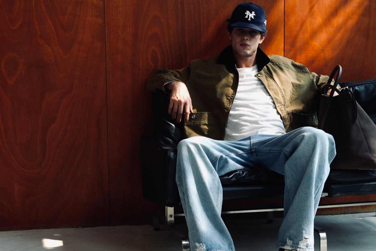 A man sits on a black leather lounge chair against a warm wood-paneled wall, wearing a dark navy baseball cap with a white logo, a brown jacket over a white t-shirt, light blue jeans, and dark sneakers with yellow laces. A dark brown tote bag rests on the chair beside him.